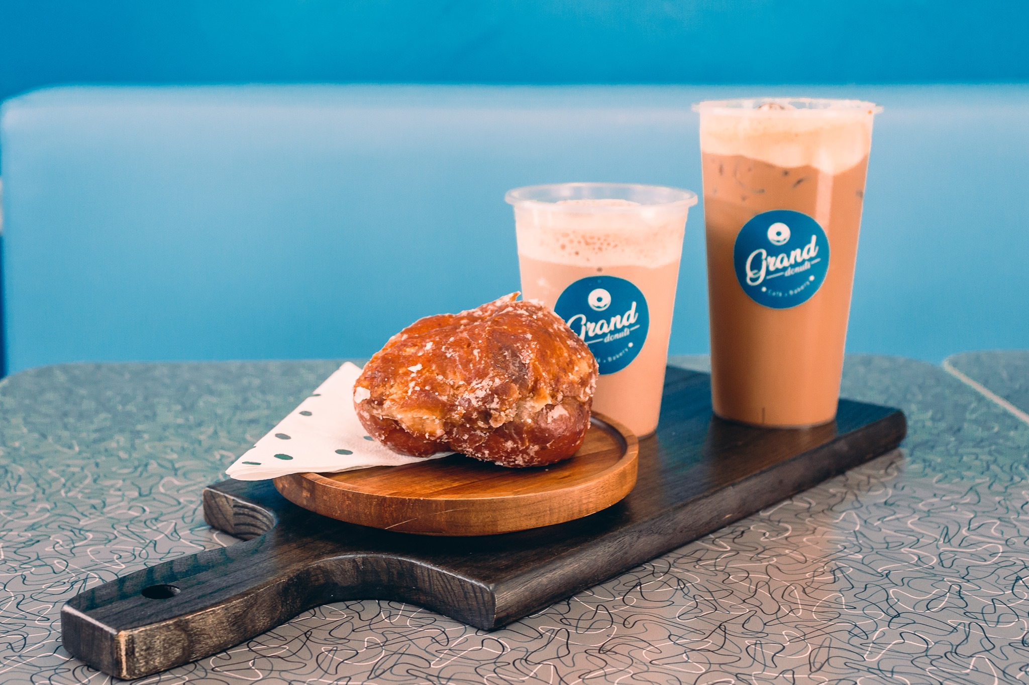 Fresh apple fritter on a wooden board with two branded GrandDonut iced drinks