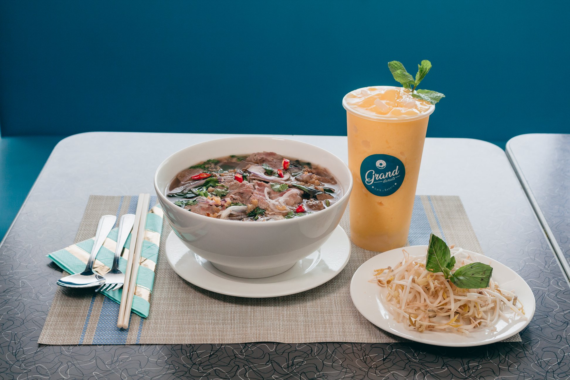 Bowl of beef pho with branded GrandDonut drink and bean sprouts on teal-accented table