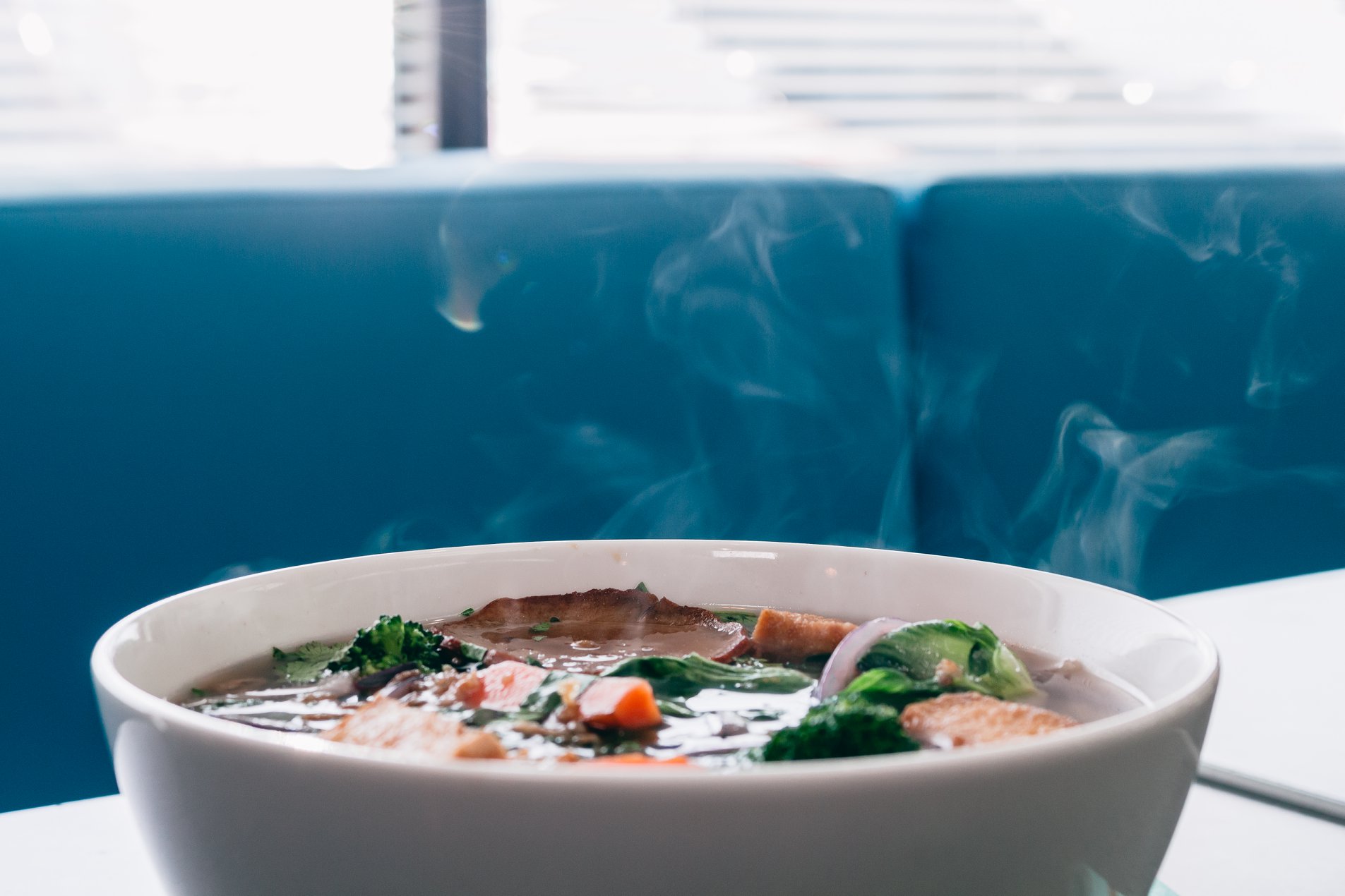 Steaming bowl of fresh pho with vegetables against teal booth seating at GrandDonut Cafe