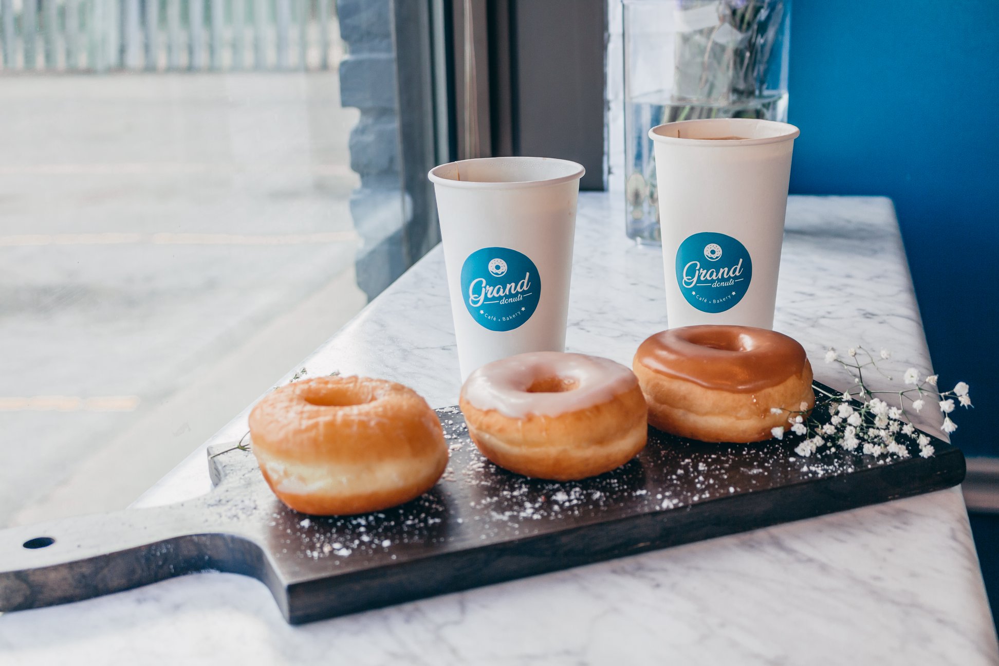 Three glazed donuts on a dark wooden board with branded GrandDonut coffee cups and baby's breath flowers