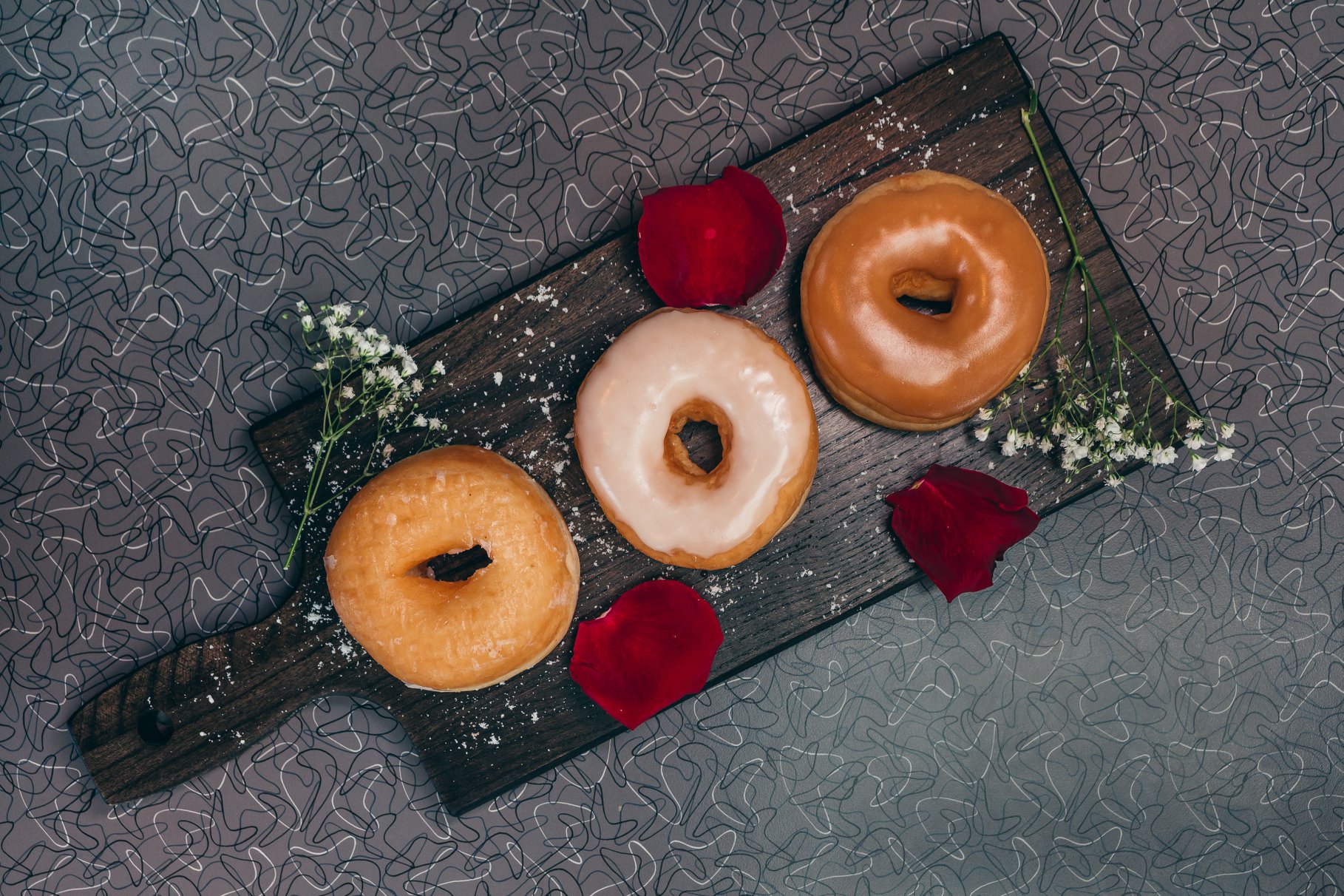 Three artisan donuts on a dark wooden board with rose petals and baby's breath, overhead view